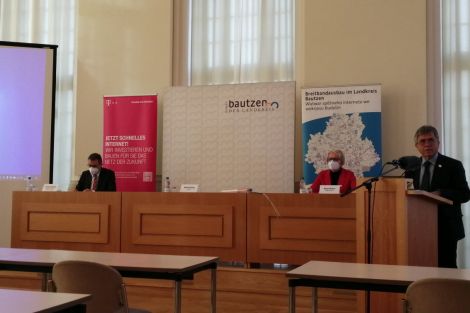 Pressekonferenz, 2 Männer und 1 Frau. Ein Mann redet am Podest. Im Hintergrund wird eine Powerpointpräsentation gezeigt.